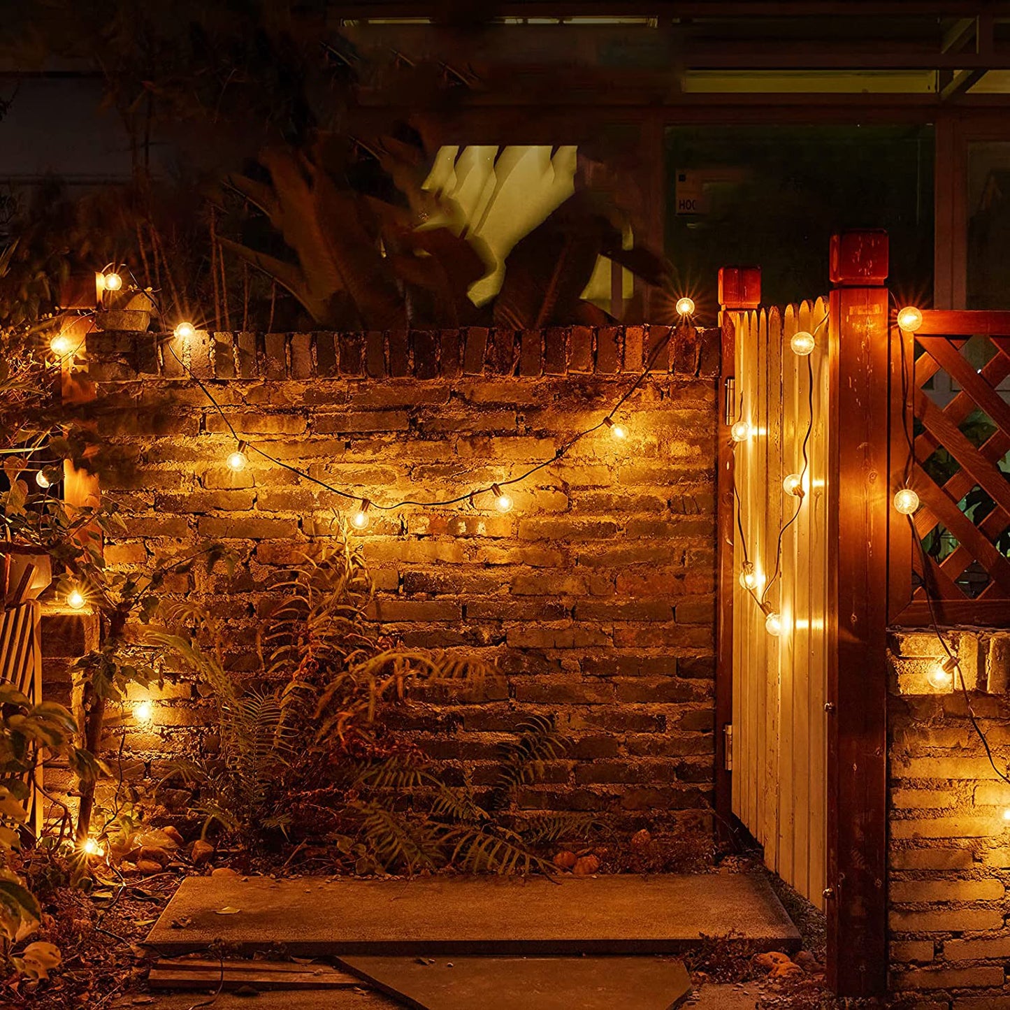 Warm yellow camping lights stringed around brick wall and wooden fence creating cozy outdoor lighting