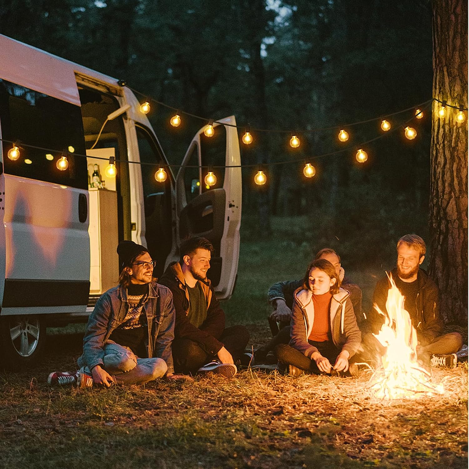 Group of friends sitting by a campfire near a van with warm led outdoor lights strung above them in the forest