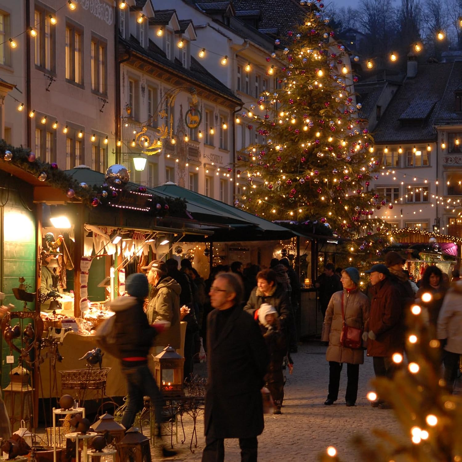 Warm glowing led outdoor lights strung above festive market stalls and a decorated Christmas tree at night